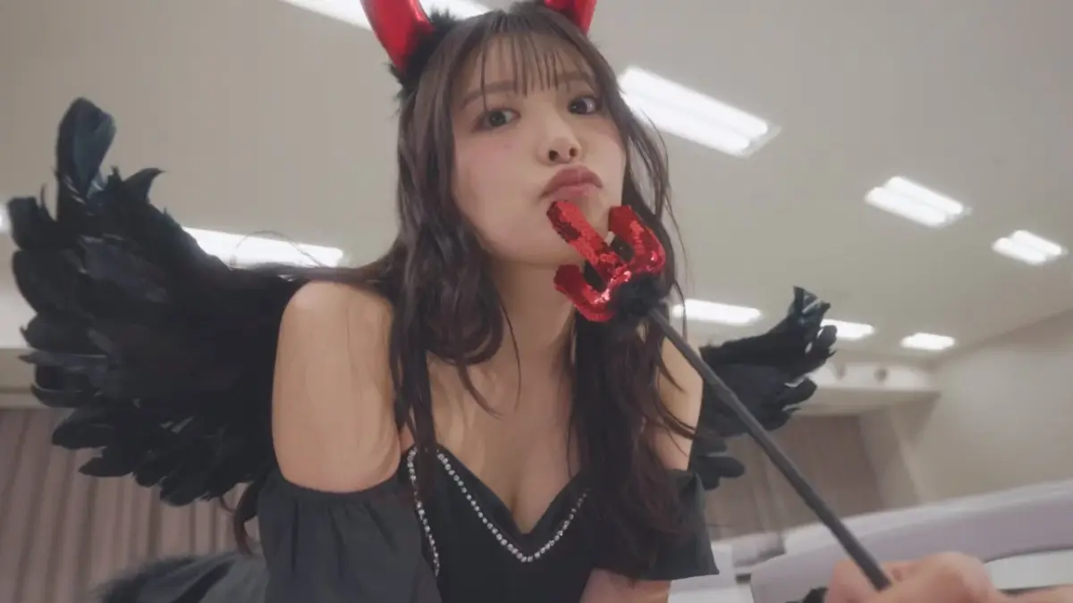 黒い悪魔のコスチュームを着た若い女性が、赤い角と黒い羽根をつけ、赤い小さな三叉の槍を口元に当ててポーズをとっています。