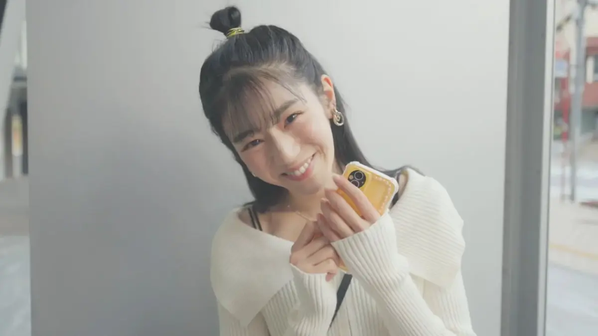 白いカーディガンを着用し、ハーフアップにした髪型の若い女性が、笑顔でスマートフォンを手にカメラを見つめているポートレート写真