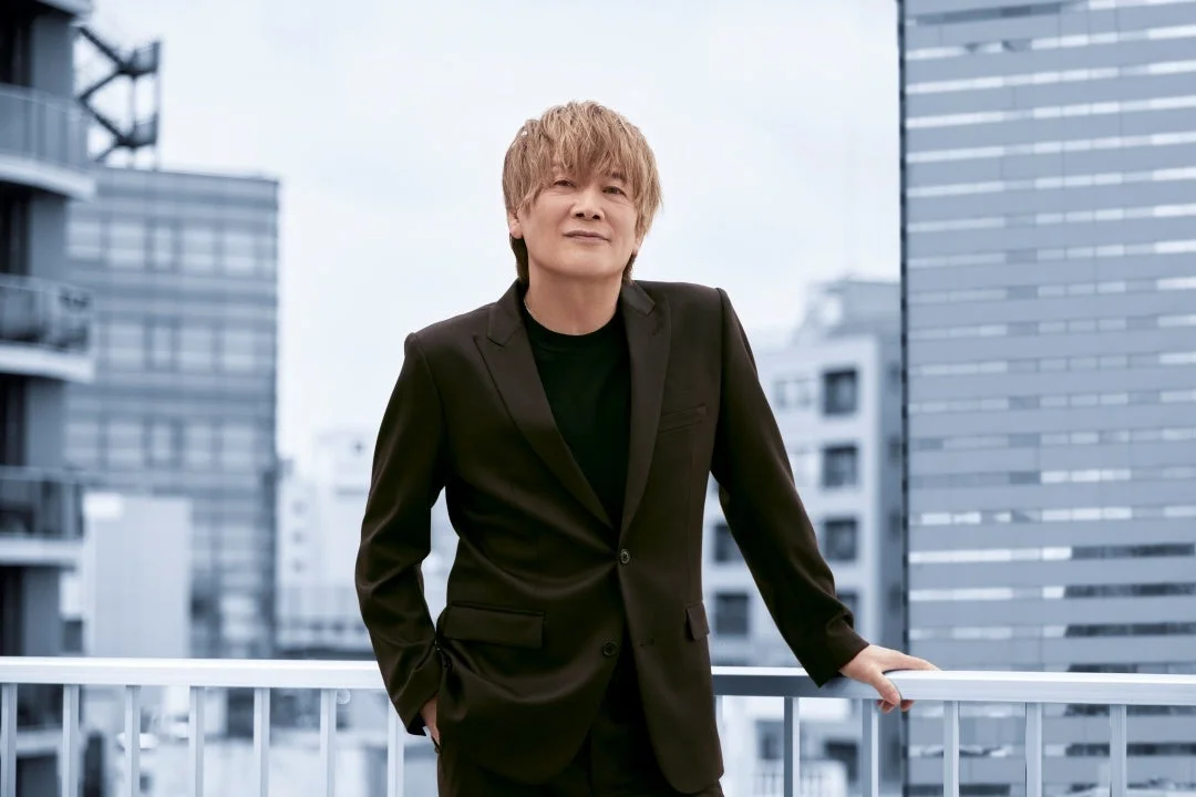 茶色の髪の男性が黒いスーツを着て、都市の建物を背景にバルコニーでポーズをとっています。