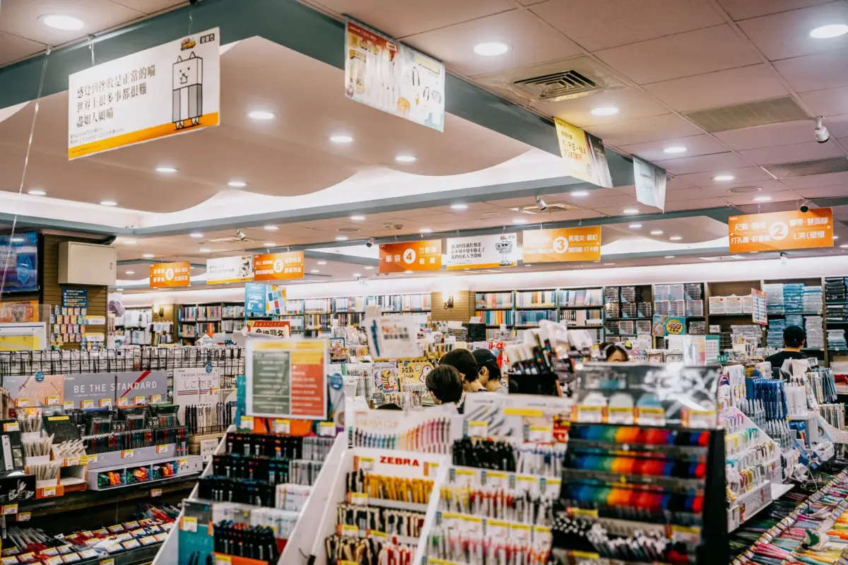 文具や画材が豊富に並ぶ広々とした小売店の店内。整然と陳列された商品棚や、買い物客の姿が見える。天井からはカテゴリーを示す看板や、可愛らしいキャラクターの励ましのメッセージボードが吊るされている。