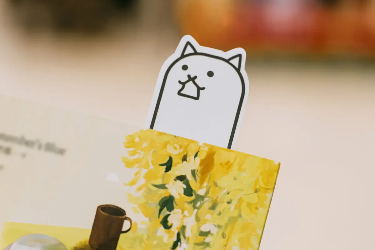 にゃんこ大戦争のキャラクターのような白い猫のブックマークが、黄色い花とマグカップのイラストが描かれた本に挟まれているクローズアップ画像です。可愛らしいデザインが特徴的です。