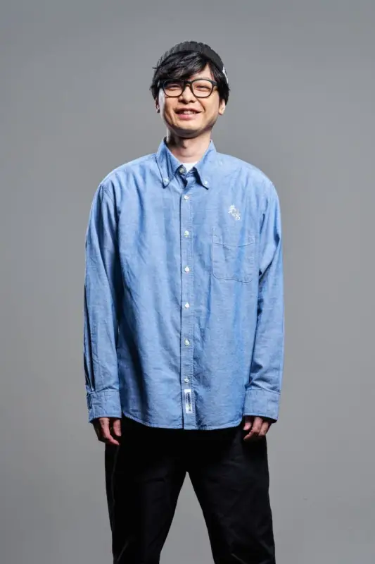 笑顔の男性が、ニット帽とメガネ、青いシャツと黒いパンツのカジュアルな服装で、シンプルな背景の前でポーズをとっているポートレート写真です。