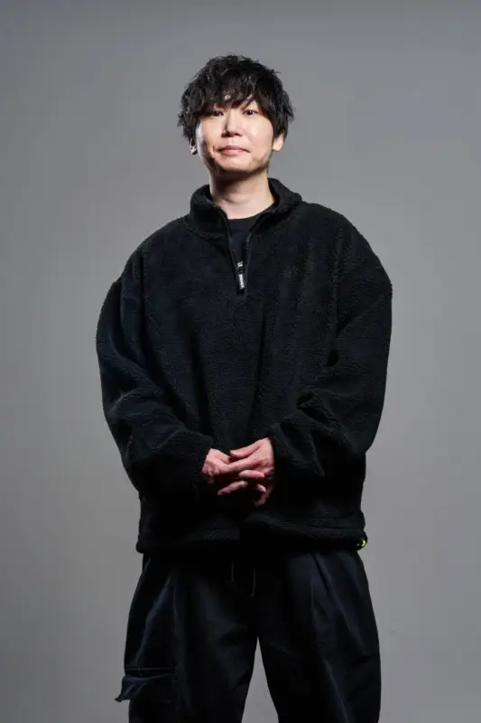 灰色の背景の前で立つ黒髪の男性。黒いフリース地のハーフジッププルオーバーと濃色のパンツを着用し、両手を組んでいる。カメラに向かって穏やかに微笑んでいる。