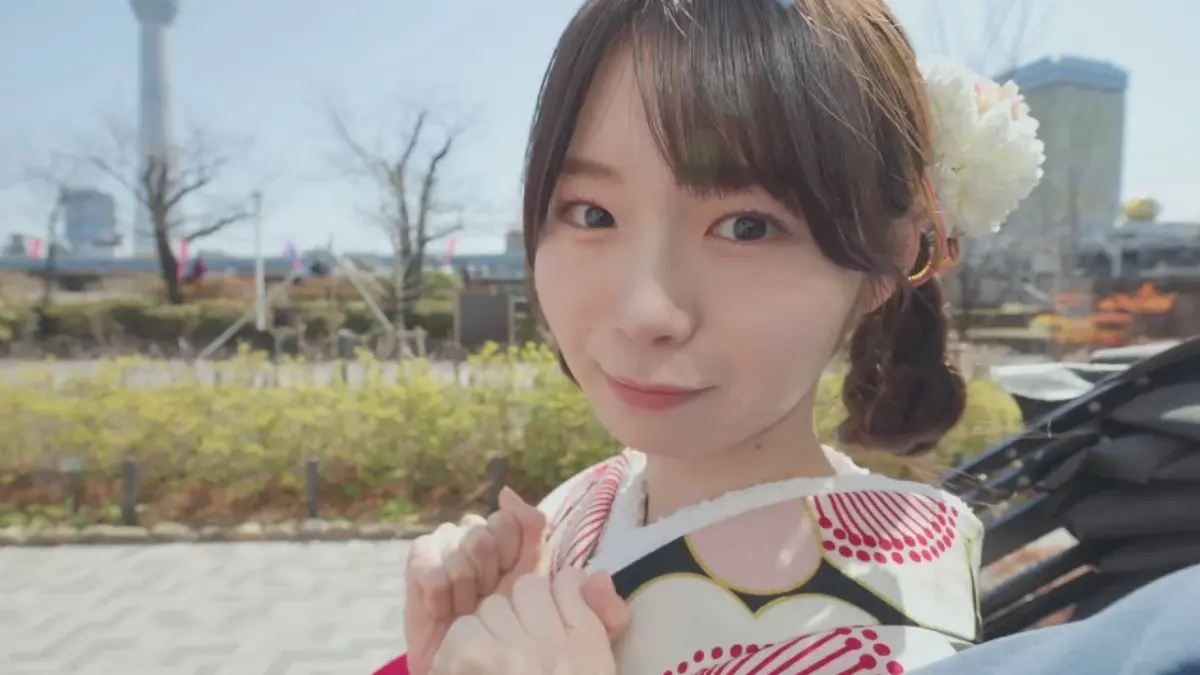 晴れた屋外で着物を着た若い女性がカメラに向かって微笑んでいます。髪には白い花飾りが付いており、背景にはぼんやりと街並みが広がっています。